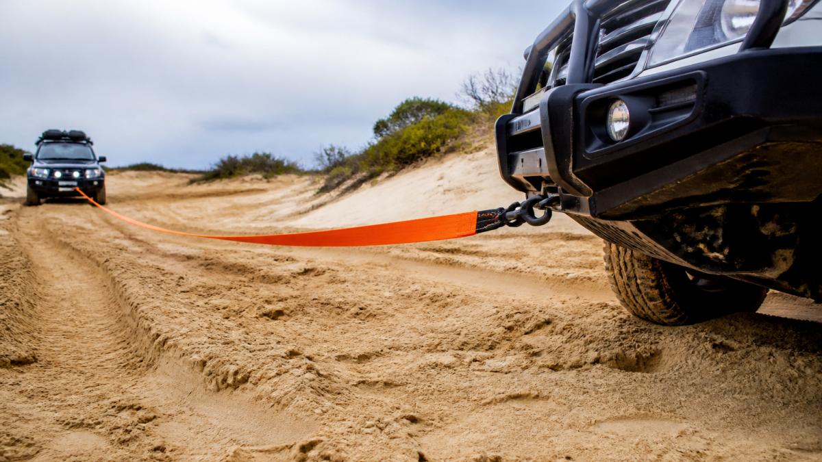 4WD vehicle with snatch strap attached to recovery points, strap under tension during recovery pull on sandy terrain, another vehicle visible in background