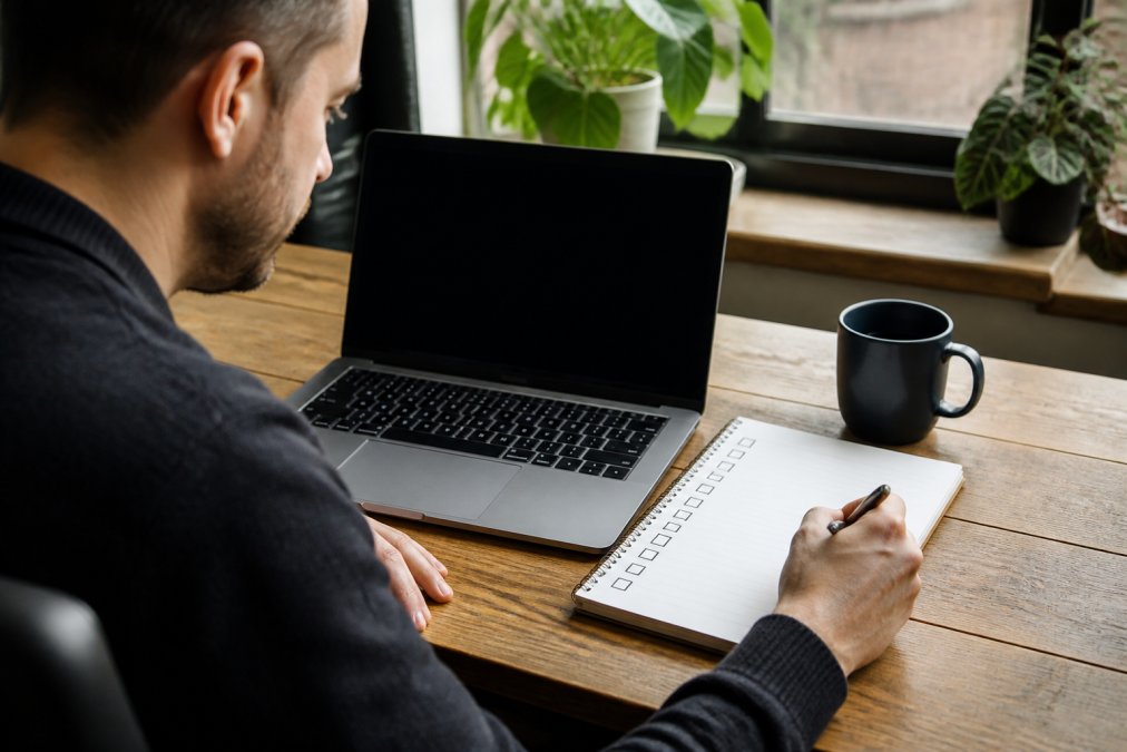 Webmaster achter bureau met laptop, notepad met checklist items zichtbaar, concentratie op optimalisatietaken