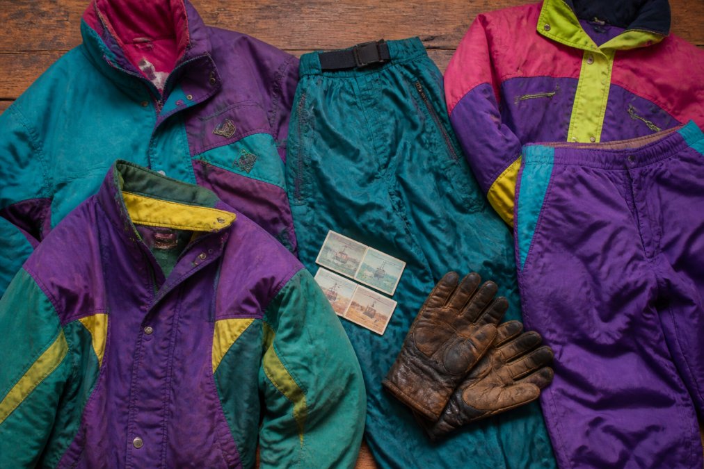 A flat-lay of colourful vintage ski jackets and retro snow pants arranged on a wooden surface, with old ski lift tickets and worn leather gloves nearby