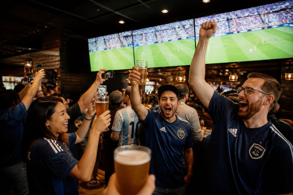 A lively group of soccer fans in team jerseys cheering and raising pints at a crowded sports bar, multiple HD screens showing a live match in the background, warm ambient lighting