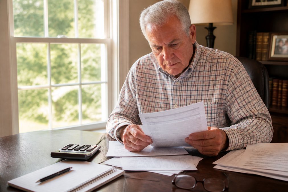 Retiree reviewing financial documents and statements at a desk with a calculator, notebook, and reading glasses nearby, natural afternoon light streaming through office window