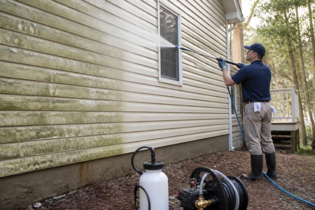 A technician in branded workwear applying a low-pressure soft wash solution to vinyl siding on a residential home, with a soft mist visible from the nozzle and green algae staining visible on the siding before treatment