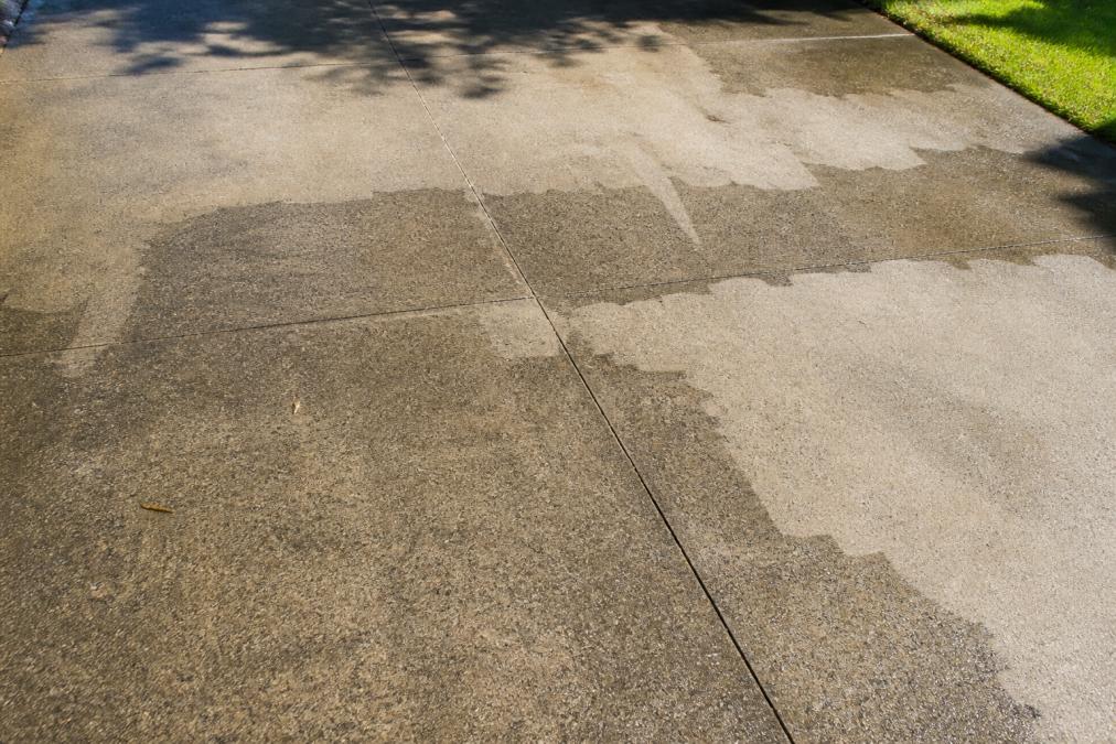 Close-up of a residential concrete driveway surface in afternoon sunlight showing clearly defined uneven patches, with darker recently cleaned areas contrasting sharply against lighter, dirtier sections that were missed or cleaned inconsistently