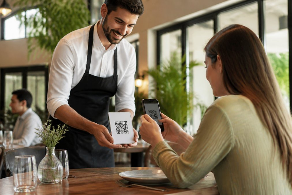 Un camarero mostrando un código QR en la mesa a un cliente que lo escanea con su móvil en un restaurante moderno y luminoso