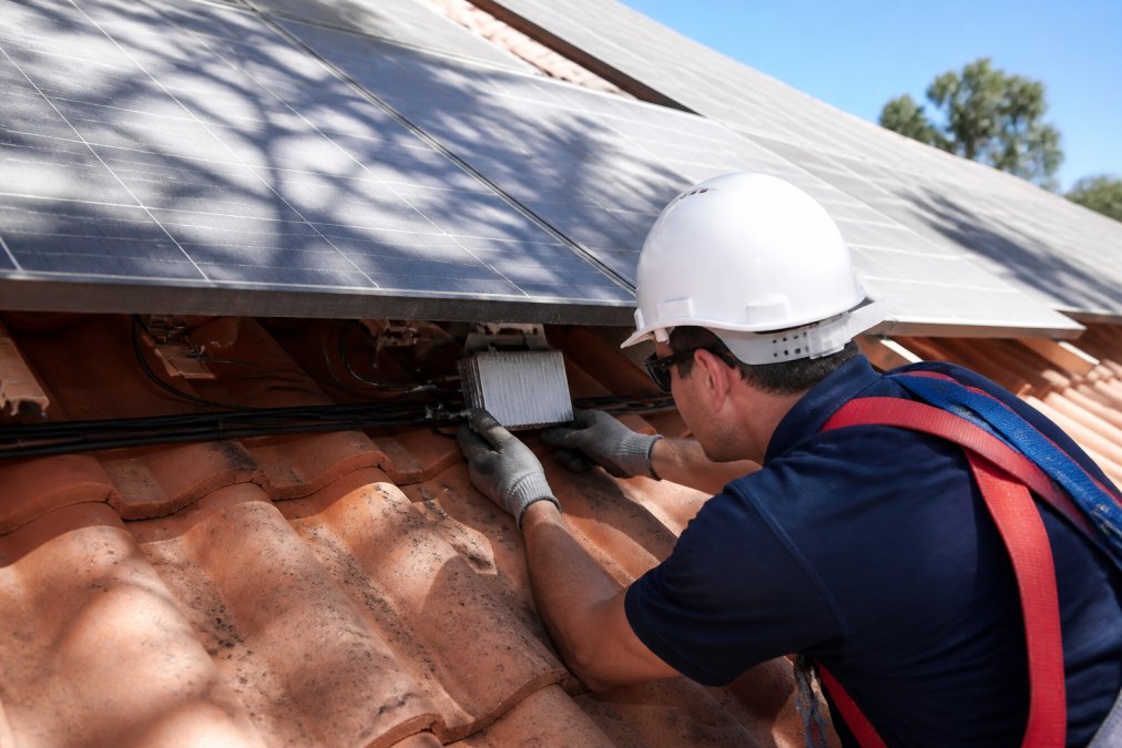A solar installer on a tiled roof securing a microinverter beneath a panel, with dappled tree shade falling across part of the array and clear sky beyond