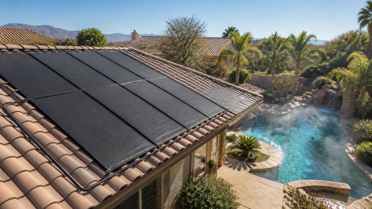 Aerial view of solar collector panels installed on a residential roof overlooking a backyard swimming pool on a sunny day, with clear blue skies and visible heat rising from the water surface
