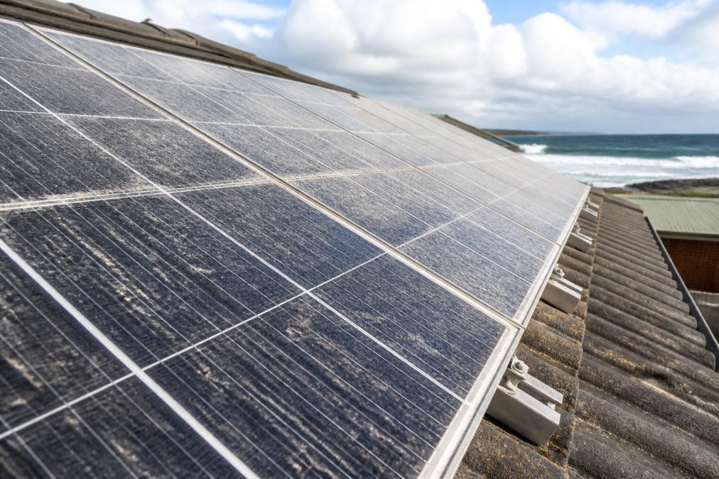 Close-up of solar panels on a residential roof with visible dust accumulation, salt spray residue, and weathering marks from coastal environment exposure, showing real-world environmental stress on module surfaces