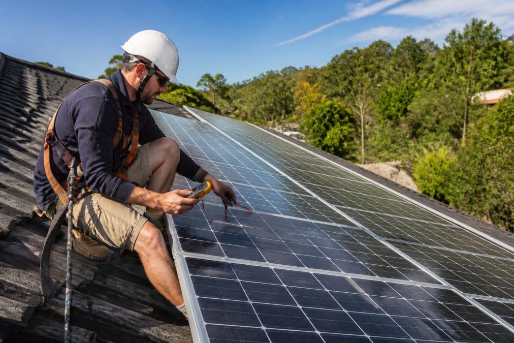 Professional technician in safety gear and harness inspecting solar panel array on residential rooftop, using handheld testing equipment to check electrical connections and module condition in bright daylight