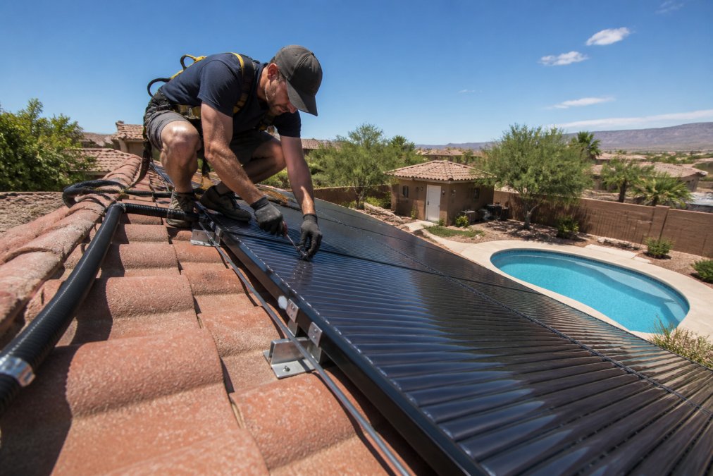 A licensed contractor on a residential rooftop carefully securing solar pool heating panels to stainless steel mounting hardware, with a clear blue sky overhead and a backyard swimming pool visible in the yard below