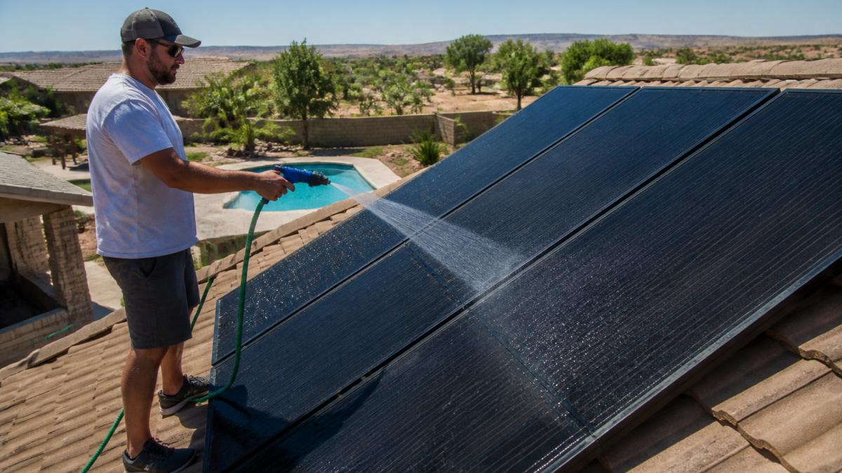 Homeowner rinsing dust and debris from roof-mounted solar pool heater panels with a garden hose