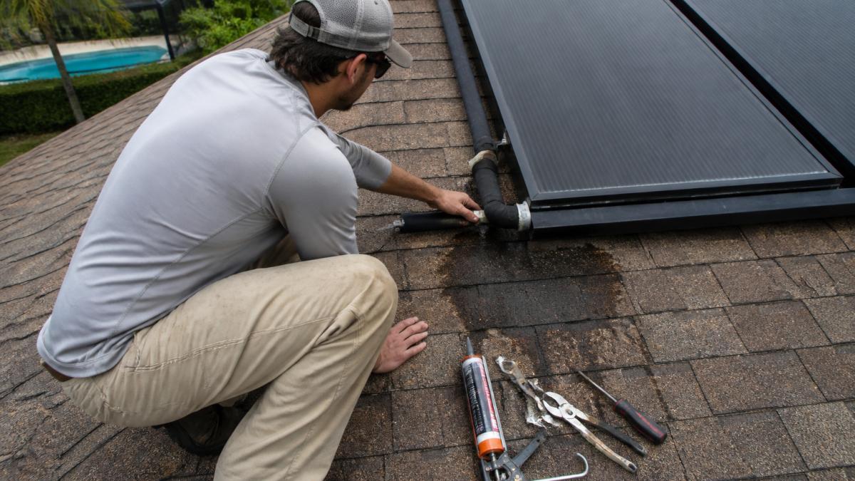 A solar pool heating technician on a shingle roof in Central Florida inspecting a row of black solar collector panels, with a wet dark stain visible on the roof decking near a panel header connection and a caulk gun and hand tools resting on the roof beside him