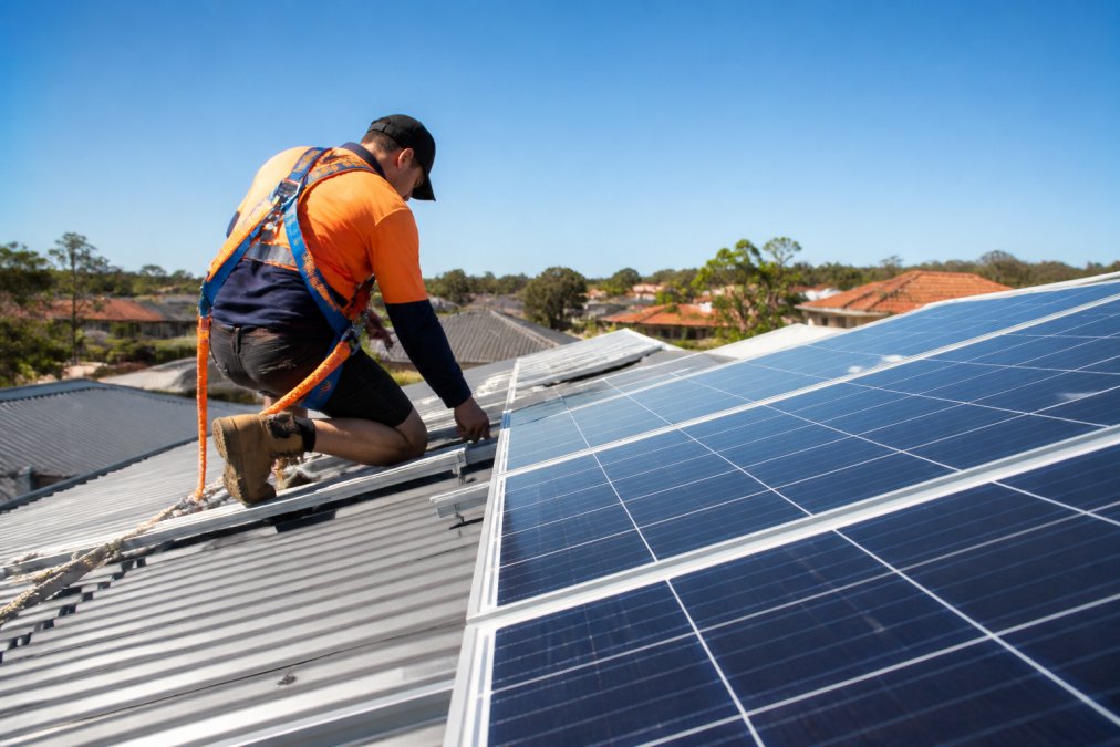 Solar installer securing photovoltaic panels to residential racking system on Australian suburban rooftop, wearing safety harness and high-visibility gear, with clear blue sky and neighbouring homes visible