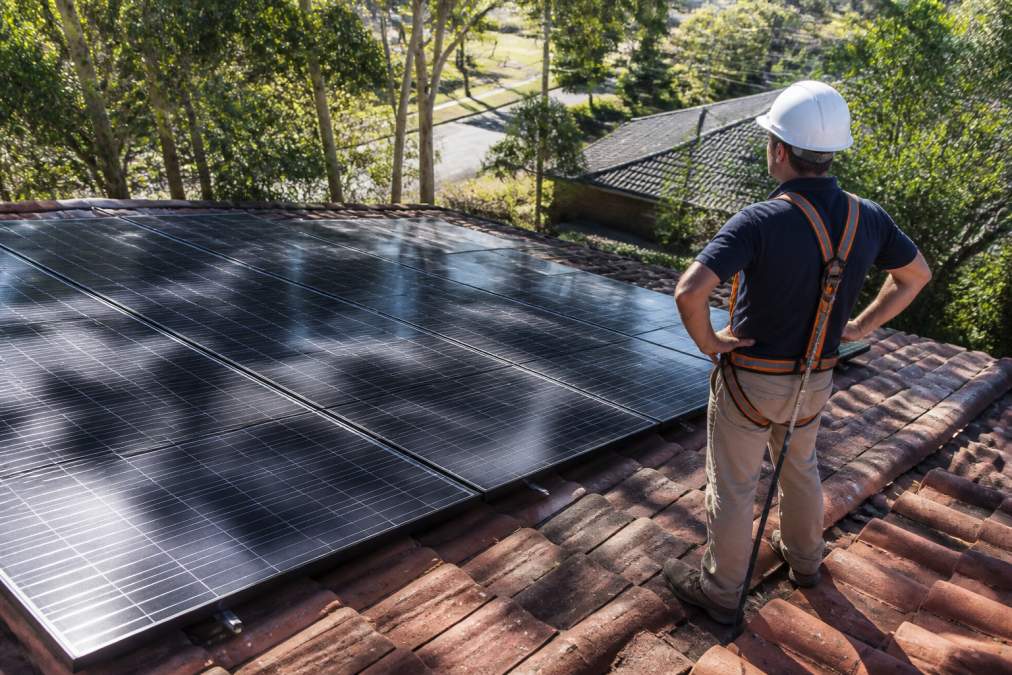 Homeowner standing on residential roof in afternoon light, with dappled shadows from nearby trees visible across solar panels, showing mix of bright and shaded areas on the roof surface