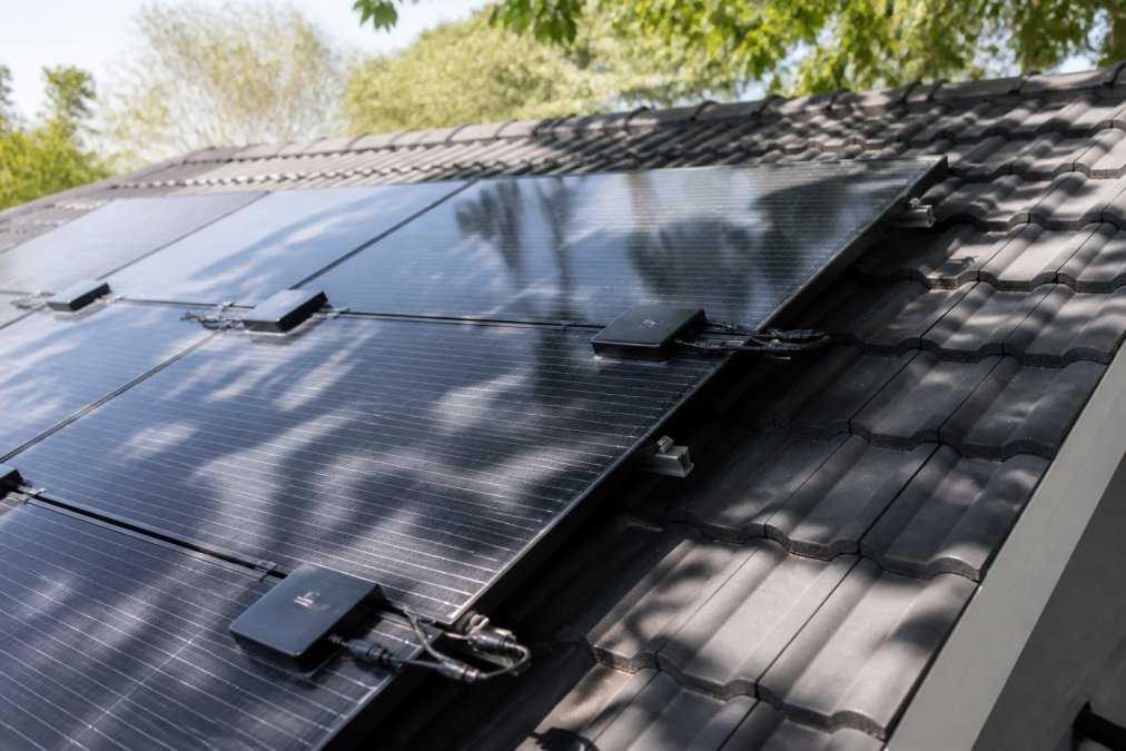 Close-up view of solar panels installed on residential roof with partial tree shade visible across the surface, showing individual microinverters mounted on panels and monitoring cables, with dappled afternoon shadows