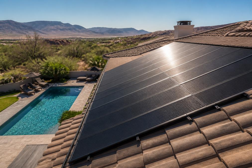 Rooftop view of solar thermal collector panels mounted on a residential home with blue pool water visible below and desert landscape in background, afternoon sunlight reflecting off the panels