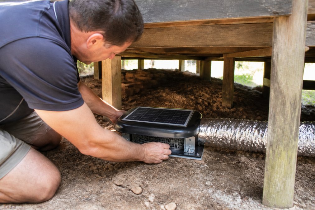 Homeowner installing a solar ventilator unit on a subfloor vent opening, showing proper positioning with mounting brackets and ducting connections in natural daylight