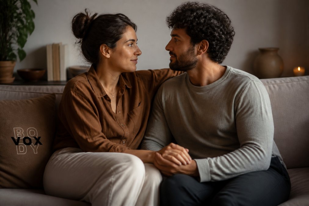 A couple sitting close together on a couch, maintaining eye contact and gentle physical contact, in a warm, softly lit living room setting