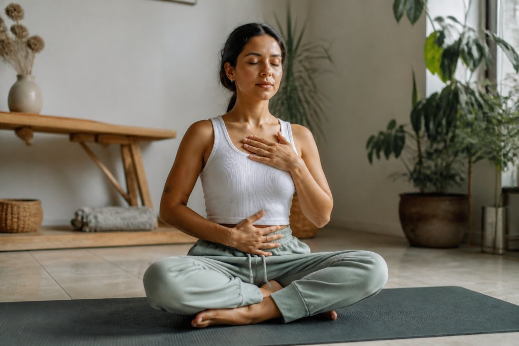 A person sitting cross-legged on a yoga mat in a sunlit room, eyes closed, one hand resting on their belly and the other on their chest, practicing slow breathing