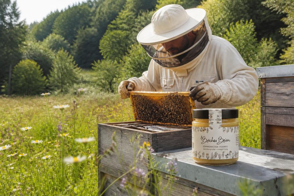 Imker bei der Arbeit an Bienenstöcken in einer luxemburgischen Landschaft, grüne Wiesen mit Wildblüten und Bäumen im Hintergrund, handwerkliche Tätigkeit beim Öffnen einer Wabe