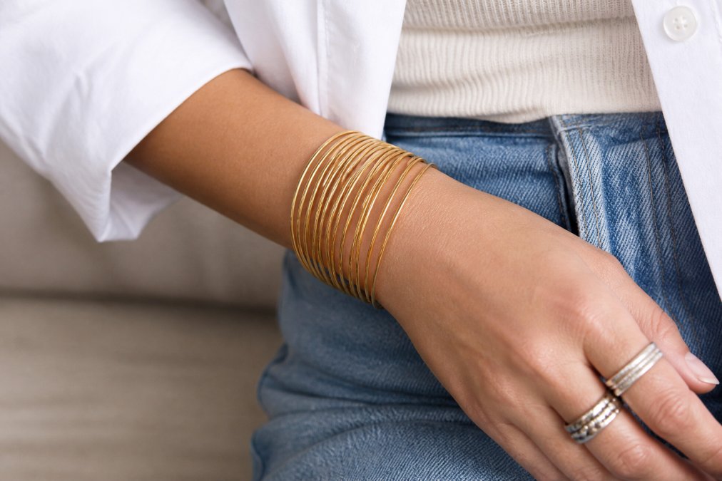 Woman's wrist and hand showing layered gold plated bracelets and bangles styled with silver rings, casual everyday outfit with natural lighting