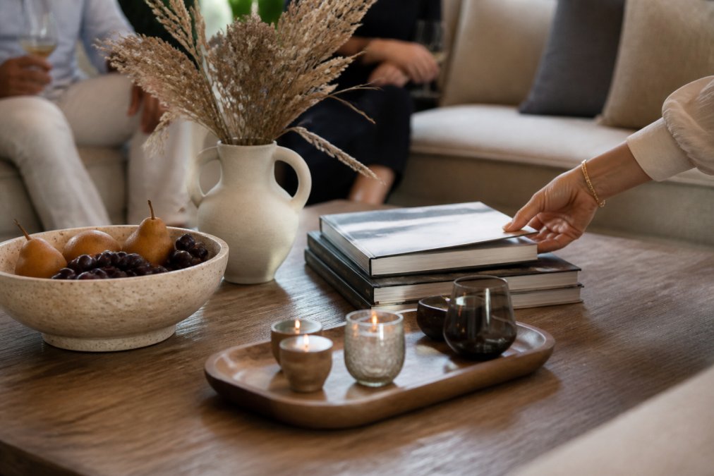 A styled coffee table displaying a travertine decorative bowl filled with seasonal fruit, a sculptural ceramic vase with dried grasses, stacked design books, and a natural wood tray holding small candles, with a guest's hand reaching for a book during an afternoon gathering