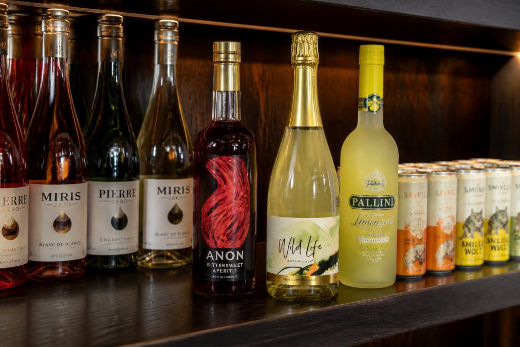 Elegantly arranged bottles of premium non-alcoholic spirits displayed on a dark wooden bar shelf with warm ambient lighting highlighting the distinctive labels and rich amber, clear, and deep coloured liquids