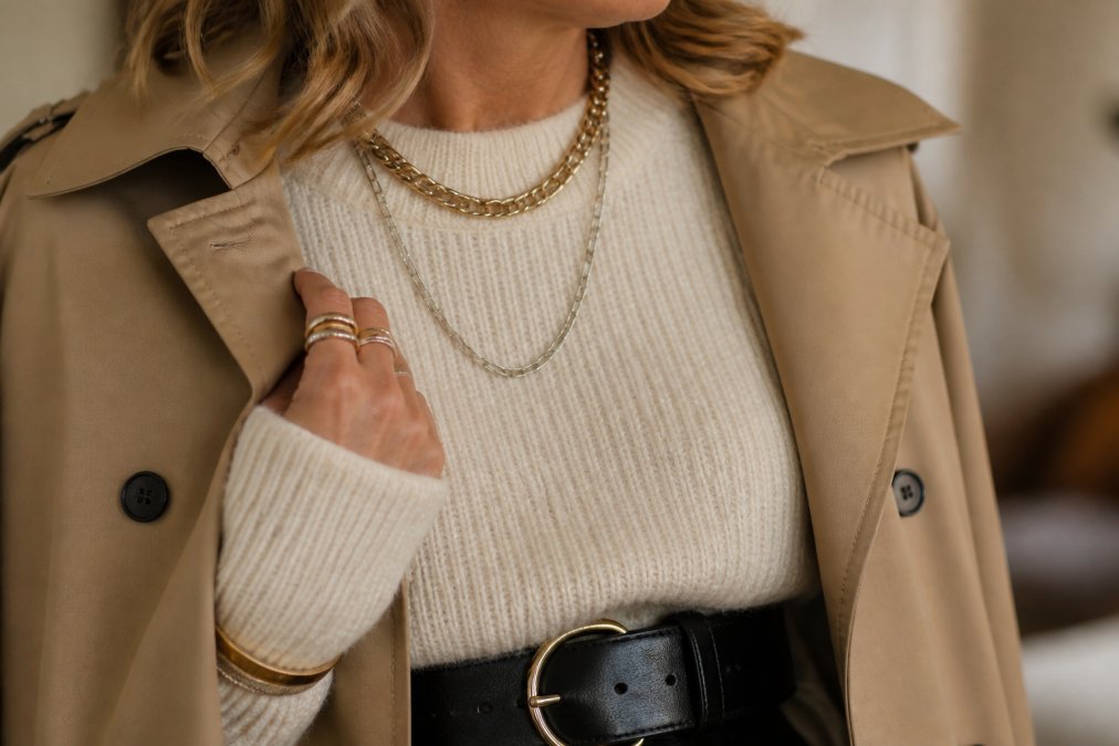 Close-up of layered textures showing a woman's outfit: cream wool sweater, camel trench coat fabric, black leather belt detail, and mixed metal jewelry catching soft indoor light