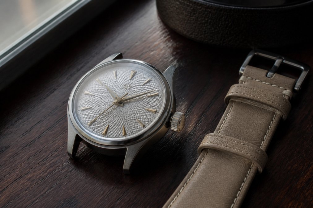 Close-up of a hand-finished titanium watch resting on a dark wooden surface beside a sand-colored leather strap, with soft natural window light highlighting the ornate guilloche dial texture and applied hour markers