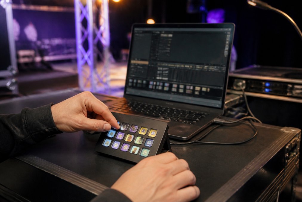 Professional operator's hands positioned over Elgato Stream Deck with illuminated scene buttons, laptop displaying DMX lighting dashboard in background during live performance setup