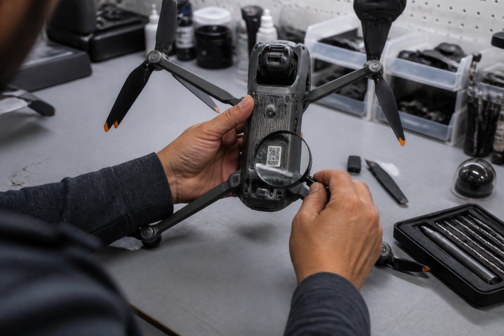 Technician's hands holding a drone while examining the serial number label on the underside with a magnifying glass, pointing to firmware version sticker, bright overhead LED lighting in workshop setting