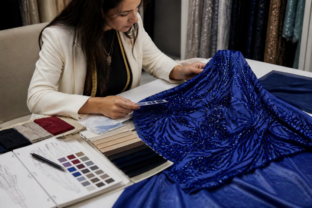 Fashion designer examining fabric swatches and samples at a workspace with a detailed checklist, color palette reference, and quality assessment notes visible on the table