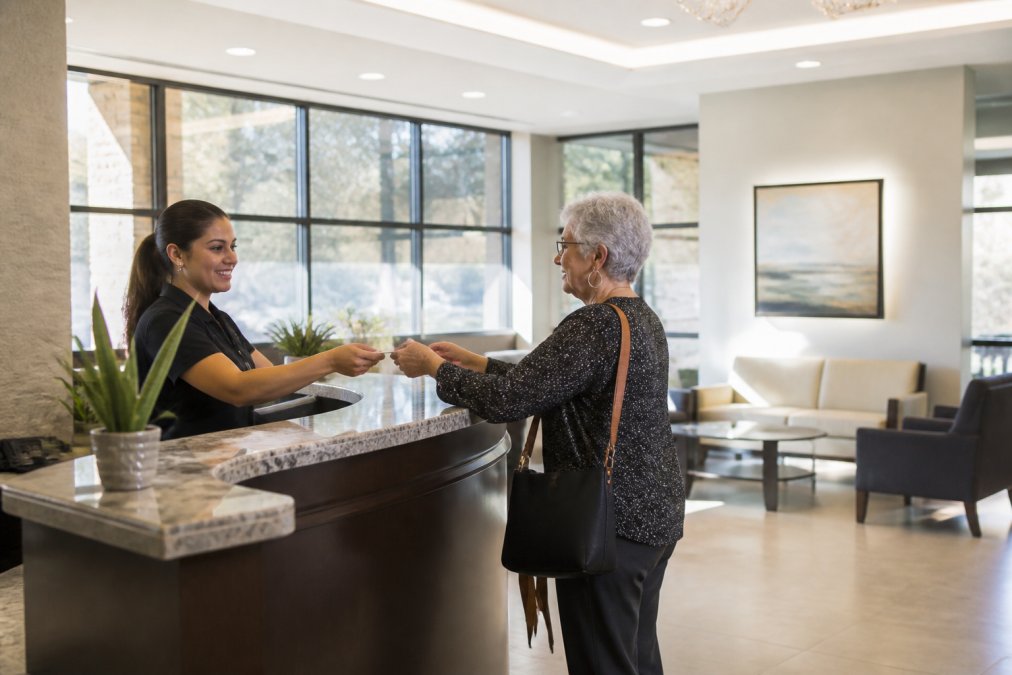 A modern eye clinic reception area with a friendly front desk staff member assisting an older patient, bright natural light through large windows, comfortable seating