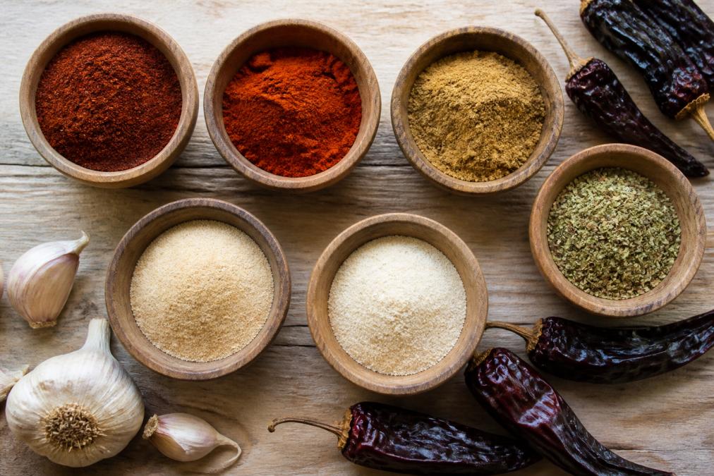 Overhead flat lay of individual spice bowls arranged on a wooden surface showing ancho chili powder, smoked paprika, ground cumin, dried oregano, garlic powder, and onion powder with fresh garlic and dried chiles nearby on a light wooden table with natural window light