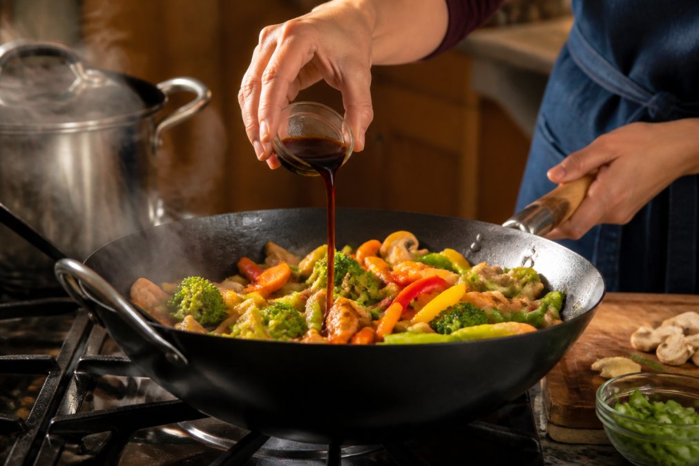 A home cook's hands drizzling coconut aminos over a colorful stir-fry in a wok with fresh vegetables, mushrooms, and steam rising, warm kitchen lighting with stainless steel cookware visible