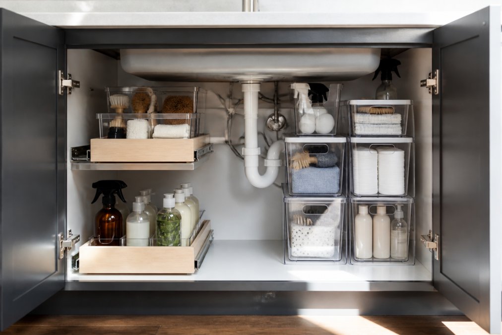 Close-up of an organized under-sink cabinet with pull-out sliding drawers, stackable waterproof bins arranged around visible U-shaped plumbing pipes, and cleaning supplies neatly sorted in labeled containers with natural lighting from above