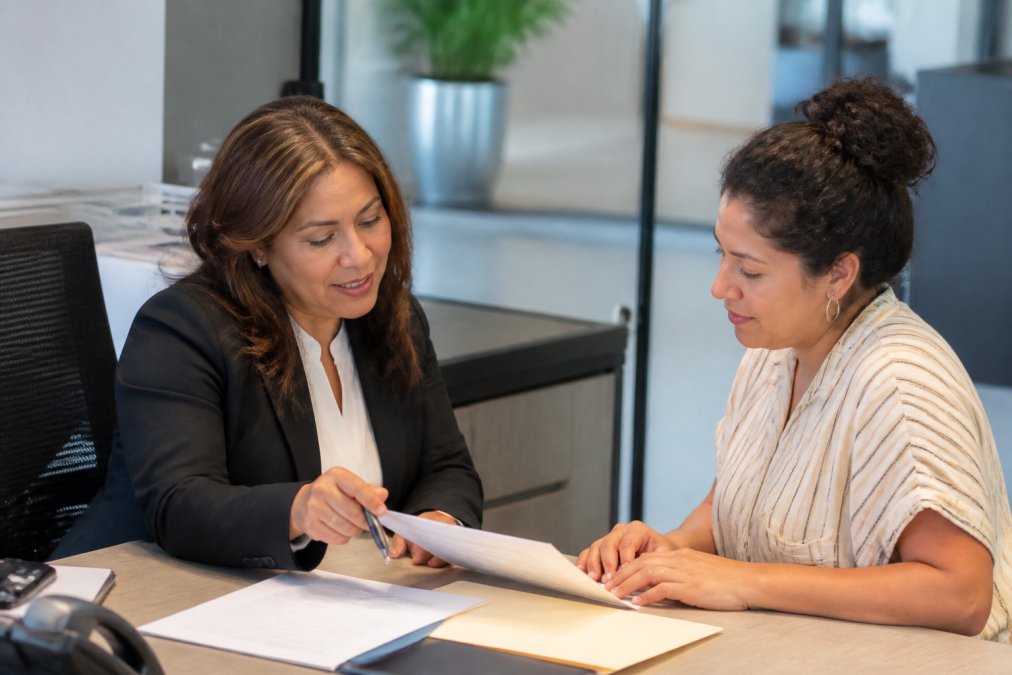 A bilingual insurance agent in professional business attire having a friendly conversation with a Spanish-speaking client in a modern office, both reviewing insurance documents together at a desk