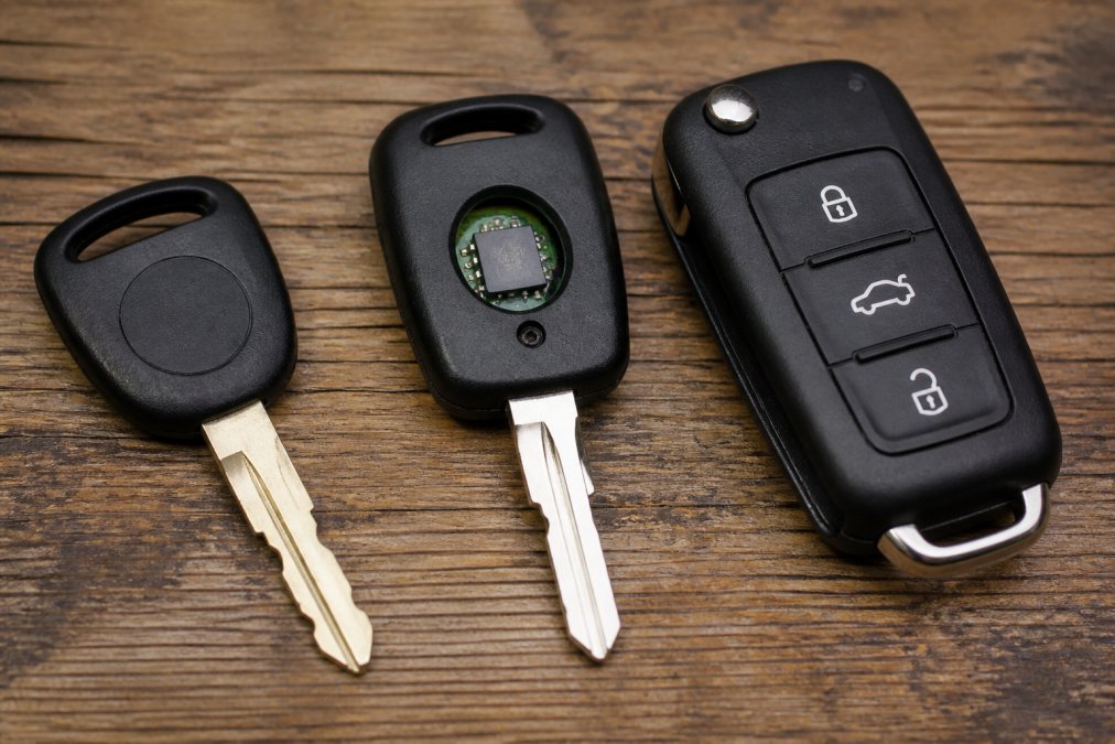 Close-up of three different car keys laid out on a wooden surface: a standard brass key blade, a transponder key with visible microchip embedded in the head, and a modern black key fob with buttons, all photographed on natural wood with soft lighting