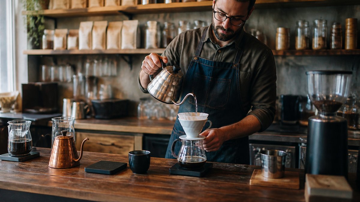 A barista behind a wooden counter pouring a pour-over coffee in a warmly lit independent café, with bags of coffee beans and brewing equipment visible on shelves behind them