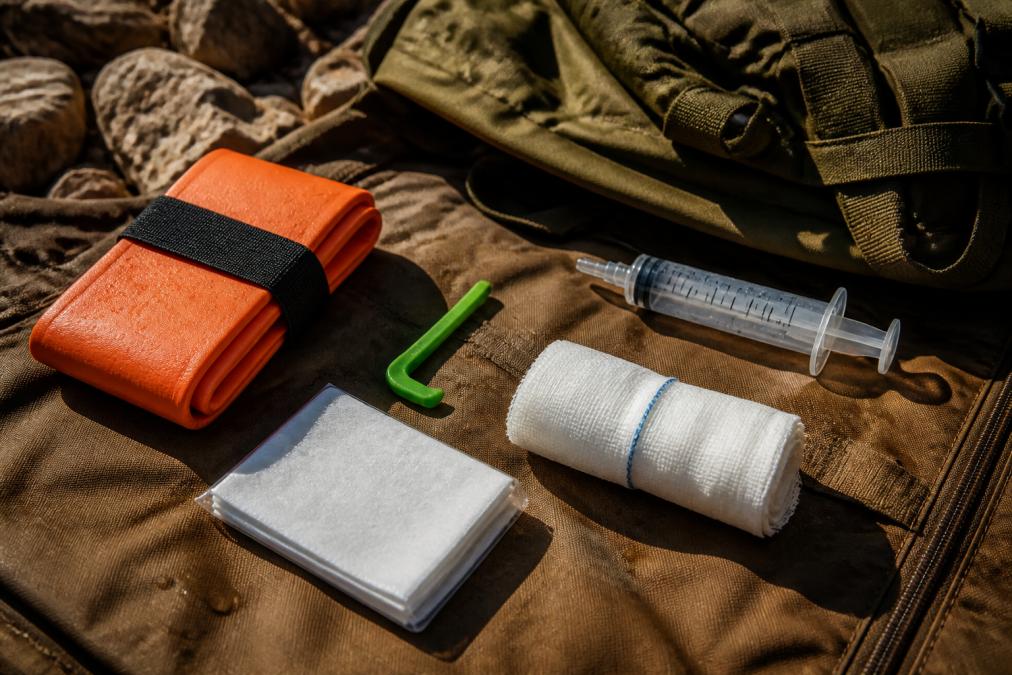 Close-up of specialized wilderness first aid supplies including SAM splint, tick twister with ID card, trauma gauze pads, and irrigation syringe arranged on a weatherproof bag with natural outdoor lighting
