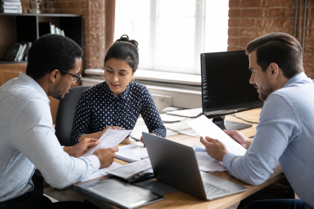 Diverse recruitment team reviewing candidate profiles and resumes at desk with multiple monitors, discussing hiring strategy with documents and laptops in modern office setting