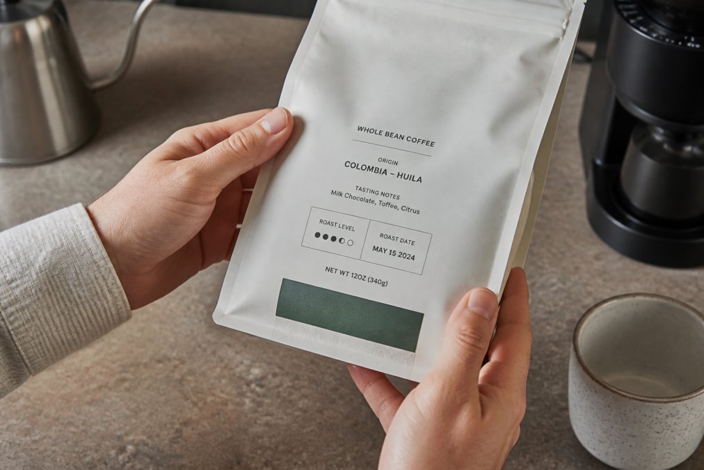 Close-up of hands holding a coffee bag label showing roast date and origin details, with a kitchen counter background