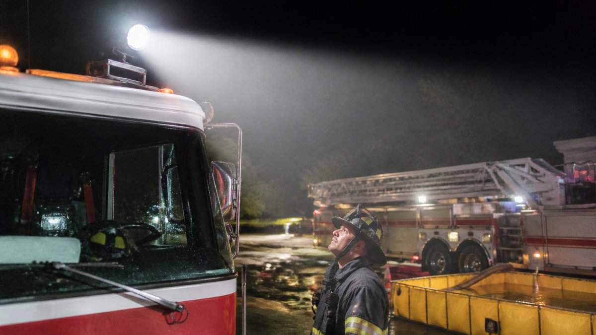 Professional firefighter standing near fire truck cab roof with mounted Spectra LED scene light illuminating a nighttime emergency work area, showing the light beam pattern across the scene