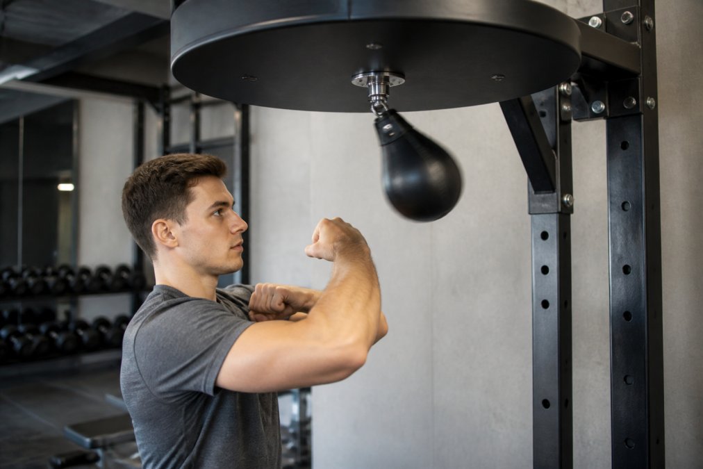Person demonstrating correct posture and arm alignment while using speed bag burn machine, showing proper shoulder stability, external rotation, and ergonomic positioning in modern fitness facility