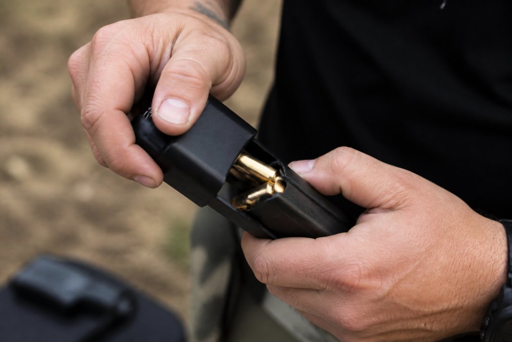 Close-up of hands loading rounds into a magazine using a speed loader at an outdoor shooting range, showing the loading mechanism in action with brass ammunition visible