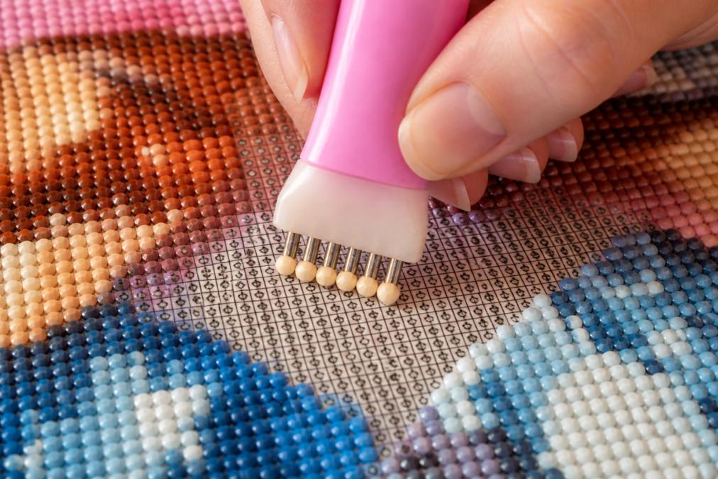 Close-up of hands using a multi-placer pen with multiple tips simultaneously placing several diamond drills onto a canvas section, showing the ergonomic grip and precise alignment of drills in a grid pattern with clear visibility of the symbols beneath