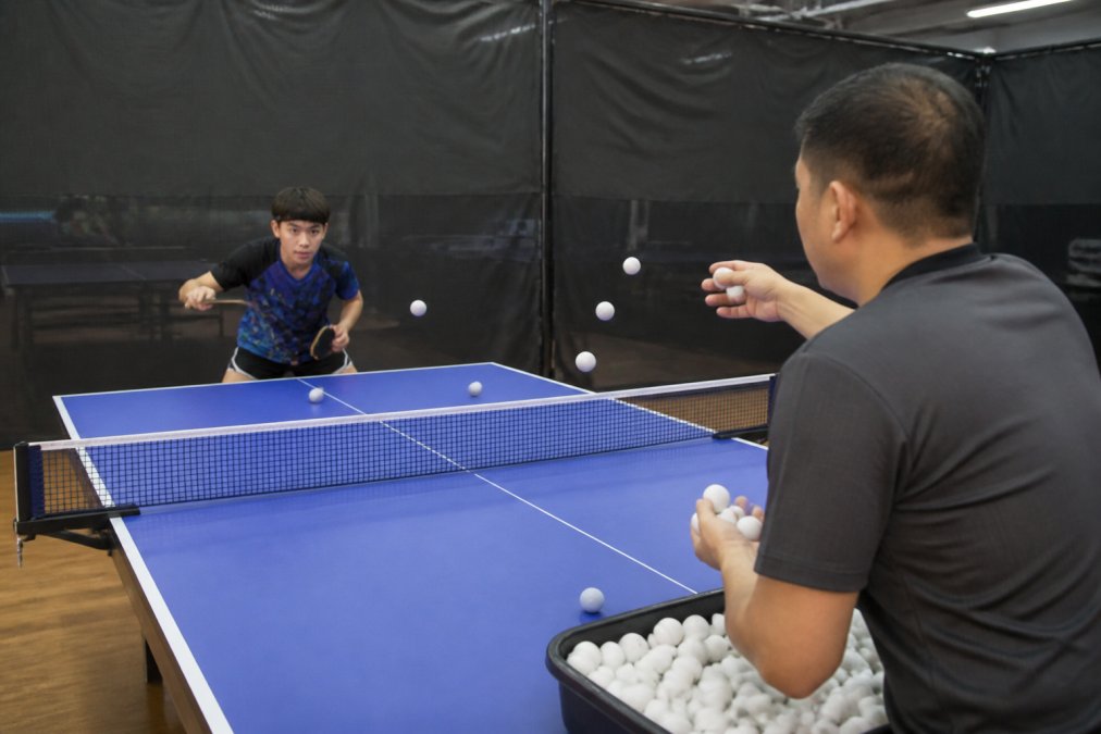 Coach feeding multiple balls to player during multi-ball drill session at table tennis table, showing rapid ball placement and player movement with focused concentration