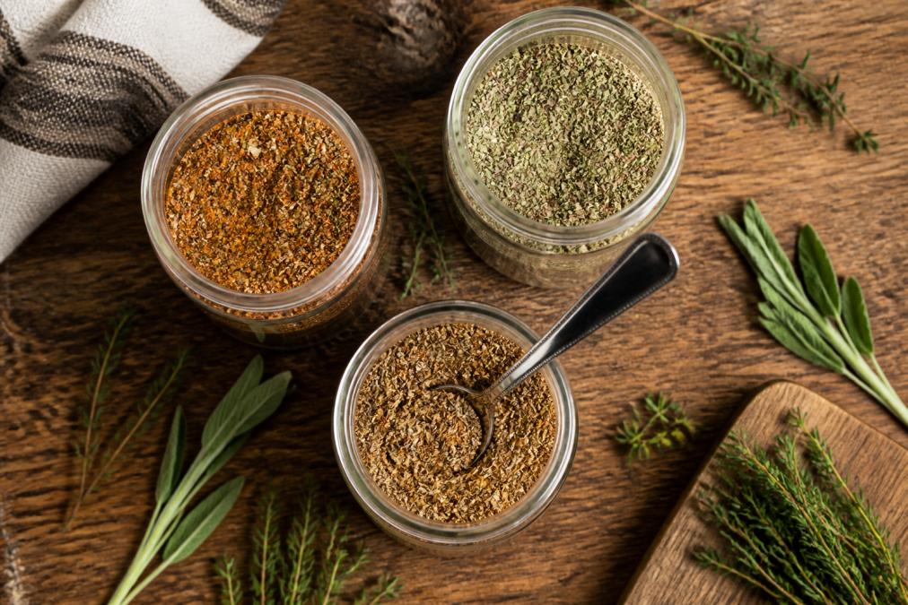 Overhead shot of three open seasoning jars on a rustic wooden kitchen counter, with a spoon resting in one jar and scattered herbs nearby, warm natural light