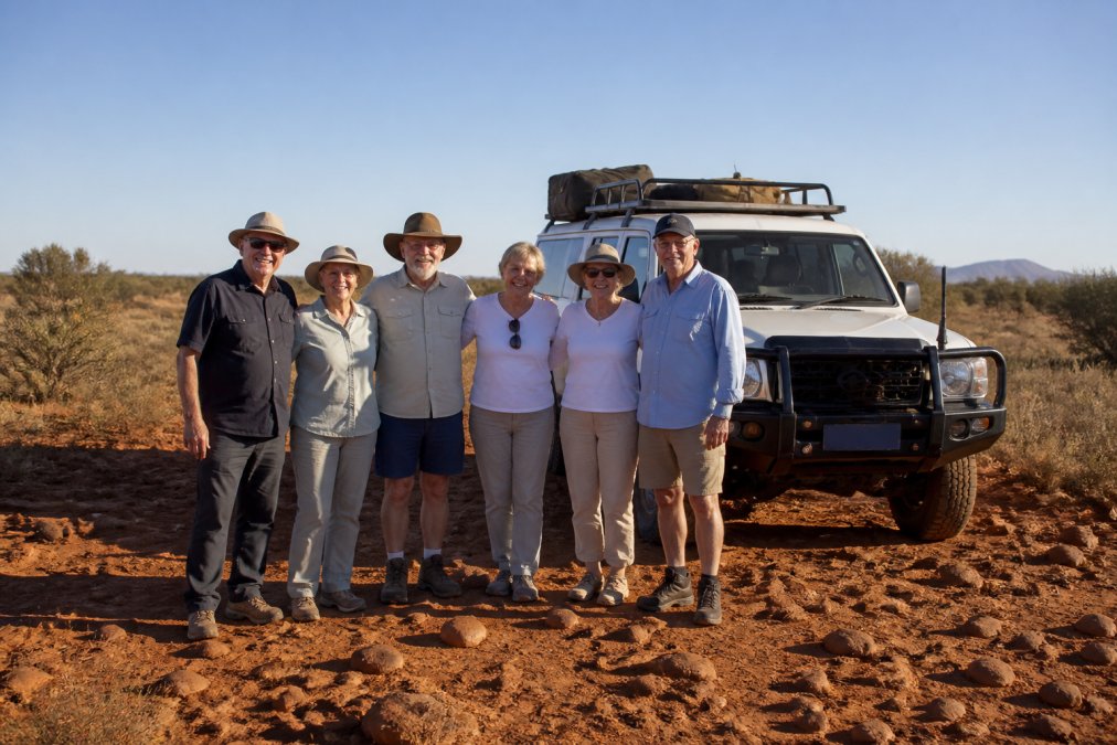 A small group of mature travellers standing together in front of a purpose-designed 4WD vehicle in the red outback landscape, smiling and relaxed, with sparse vegetation and warm afternoon light casting long shadows