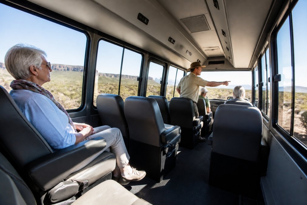 Interior view of a purpose-designed 4WD vehicle showing spacious seating with good legroom, comfortable upholstered seats, large windows positioned for viewing, and a mature traveller comfortably seated with a guide nearby pointing out landscape features