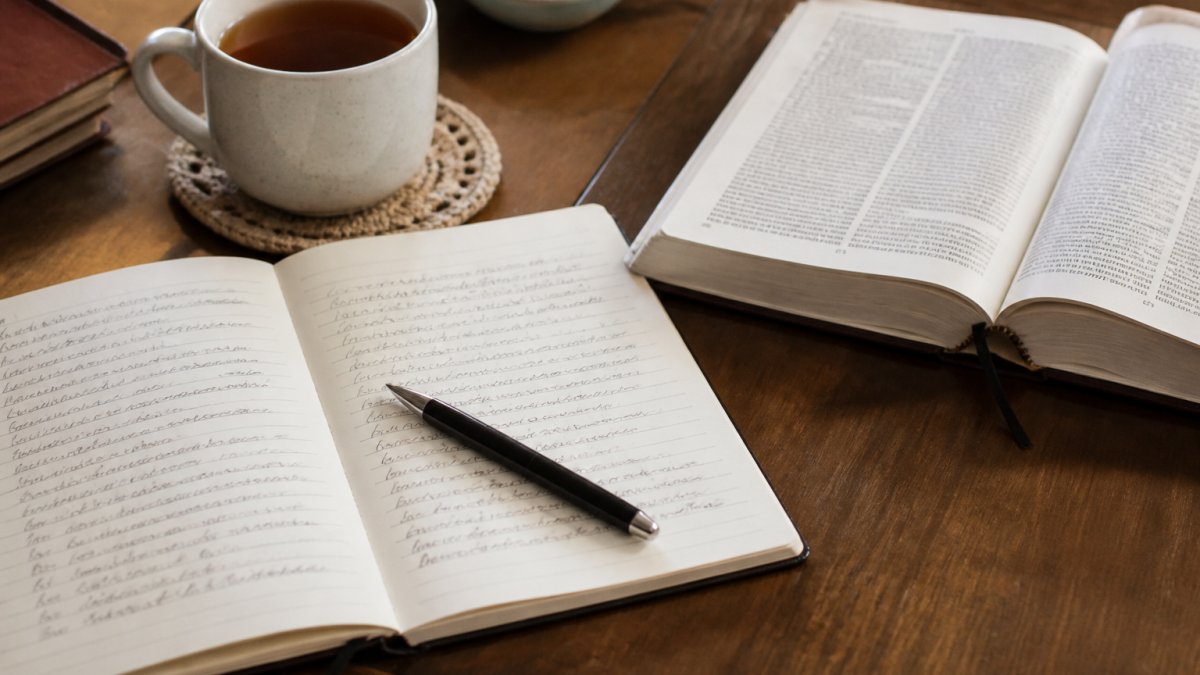 Open journal with handwritten notes, a Bible, and a pen on a wooden table beside a cup of tea